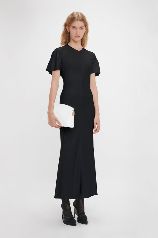 A person stands against a plain background wearing a fitted black Gathered Sleeve Midi Dress In Black by Victoria Beckham in crepe back satin and holding a white clutch. They have long hair and are wearing high-heeled shoes, adding an elegant touch with the dress's matte finish.