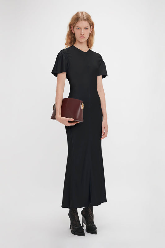 A person stands against a plain background, wearing a black dress with ruffled sleeves, black ankle boots, and holding the Victoria Clutch Bag In Burgundy Leather by Victoria Beckham.