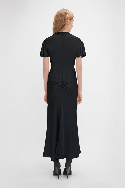 A woman with long blonde hair is standing and facing away, wearing a black Gathered Sleeve Midi Dress In Black by Victoria Beckham made from crepe back satin and black high-heeled shoes.