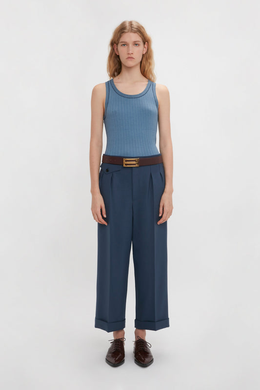A person stands facing the camera wearing a Victoria Beckham Fine Knit Micro Stripe Tank In Heritage Blue, blue wide-leg trousers, a brown belt, and black shoes. The background is plain white.