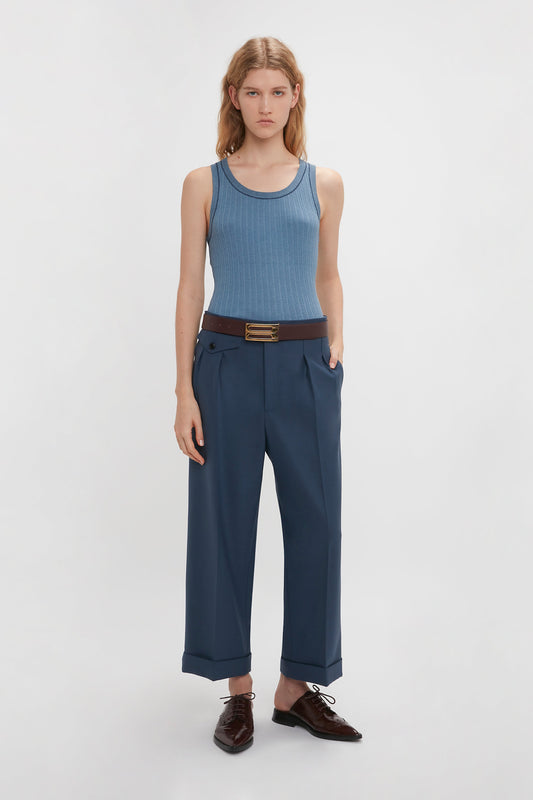 A person stands against a plain background, wearing a Fine Knit Micro Stripe Tank In Heritage Blue by Victoria Beckham, blue high-waisted pants with hands in pockets, and brown shoes.