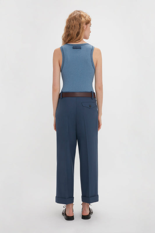 A person with wavy hair stands facing away, wearing the Victoria Beckham Fine Knit Micro Stripe Tank In Heritage Blue, dark blue belted pants with rolled-up cuffs, and black shoes.