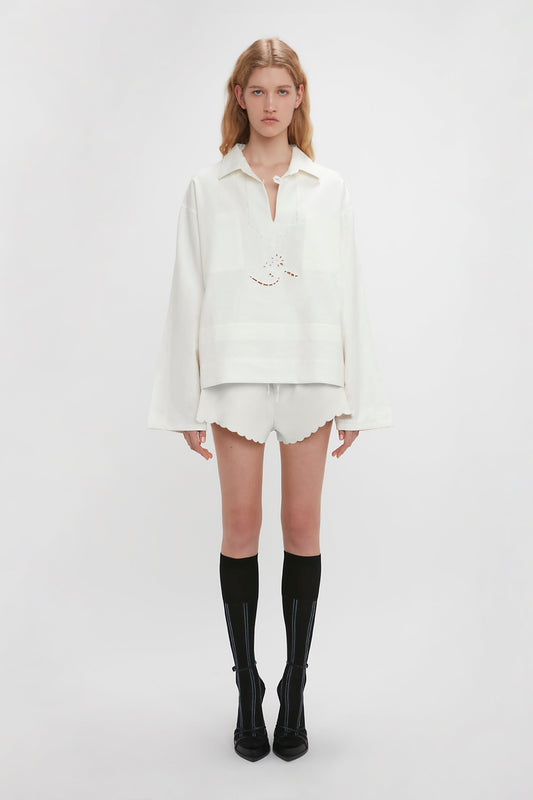 A person stands facing forward wearing an Oversized Embroidered Tunic In Antique White by Victoria Beckham and white shorts, paired with black knee-high socks and black shoes, against a plain white background.