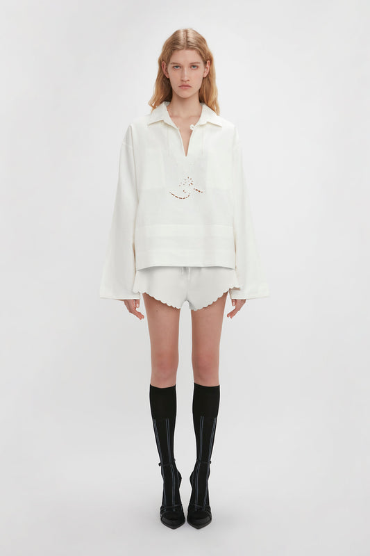 A person stands against a plain white background, wearing a loose white blouse and the Drawstring Embroidered Mini Short In Antique White by Victoria Beckham made from a breathable linen-cotton blend. Black knee-high socks paired with black shoes complete the look.