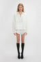 A person stands against a plain white background, wearing a loose white blouse and the Drawstring Embroidered Mini Short In Antique White by Victoria Beckham made from a breathable linen-cotton blend. Black knee-high socks paired with black shoes complete the look.