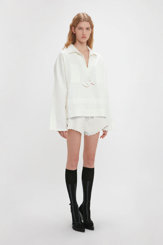 A young person with long hair is wearing a vintage-inspired, relaxed-fit Victoria Beckham Oversized Embroidered Tunic In Antique White paired with matching white shorts and knee-high black socks with white stripes. The background is plain and white.