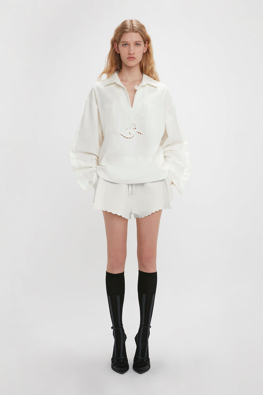 A person standing against a white background, wearing a white long-sleeve shirt with embroidered detailing, Victoria Beckham Drawstring Embroidered Mini Short In Antique White, and black knee-high socks with black shoes.