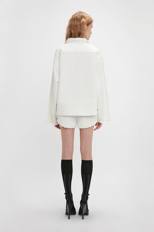 A person with long hair, dressed in an Oversized Embroidered Tunic In Antique White by Victoria Beckham and relaxed-fit tunic shorts, stands facing away from the camera. They are wearing knee-high black socks and black high-heeled shoes.