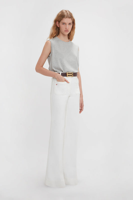 A person stands against a white background wearing a Sleeveless T-Shirt In Grey Marl by Victoria Beckham and high-waisted white pants with a dark belt.