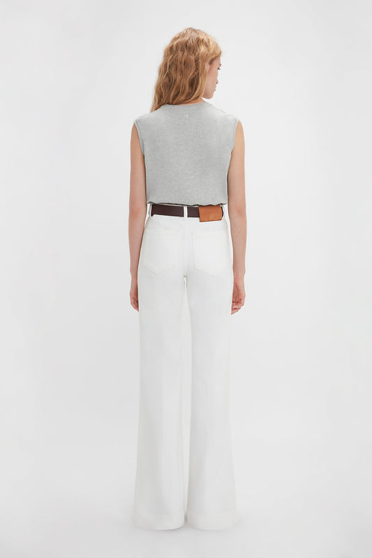 A woman stands facing away from the camera, wearing a Sleeveless T-Shirt In Grey Marl by Victoria Beckham, paired with high-waisted white wide-leg pants and a black and brown belt.
