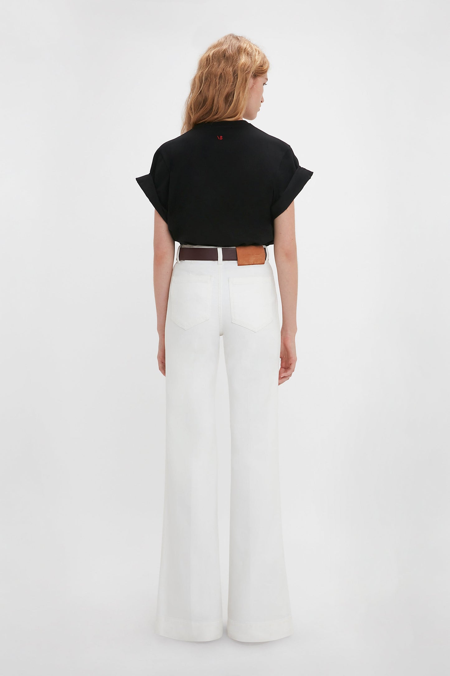 Woman in a Victoria Beckham 'Do As I Say, Not As I Do' Slogan T-Shirt in Black and white wide-leg pants, standing with her back to the camera against a plain background.