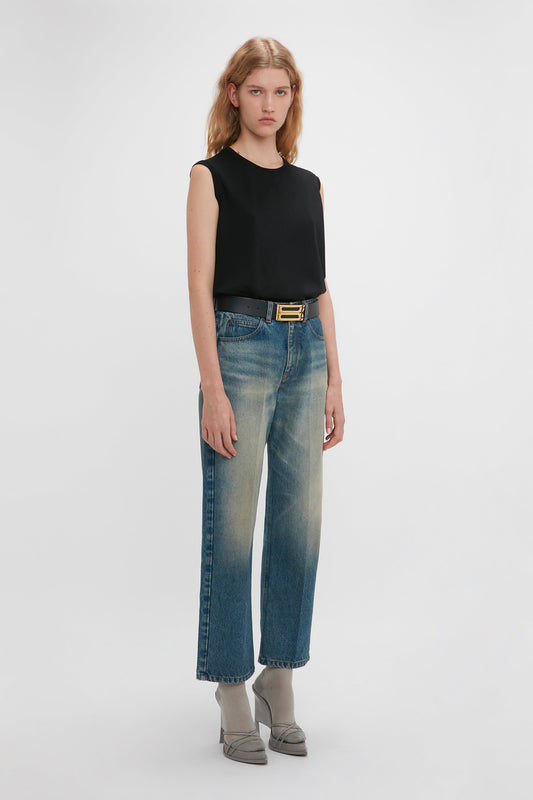 Woman in a black sleeveless top and faded blue Victoria Beckham jeans featuring a Relaxed Straight Leg Jean In Antique Indigo Wash with a gold buckle belt, standing against a white background.