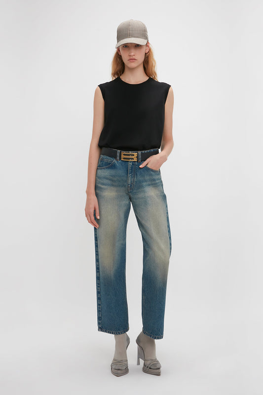Woman in black sleeveless top and faded blue jeans with a Victoria Beckham Relaxed Straight Leg Jean in Antique Indigo Wash and a brown belt, wearing a gray cap and heeled boots, standing against a white backdrop.