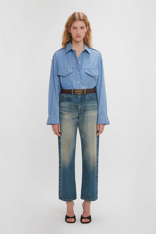 A person with long hair is standing against a plain background, wearing a Victoria Beckham Cropped Seam Detail Shirt In Steel Blue with a utilitarian sensibility, faded blue jeans, a black belt, and black open-toe shoes.