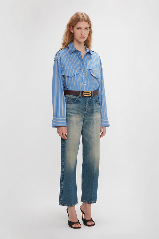 A person stands against a plain background wearing a Victoria Beckham Cropped Seam Detail Shirt in Steel Blue tucked into high-waisted faded jeans, belted with a dark belt, and black heeled sandals.