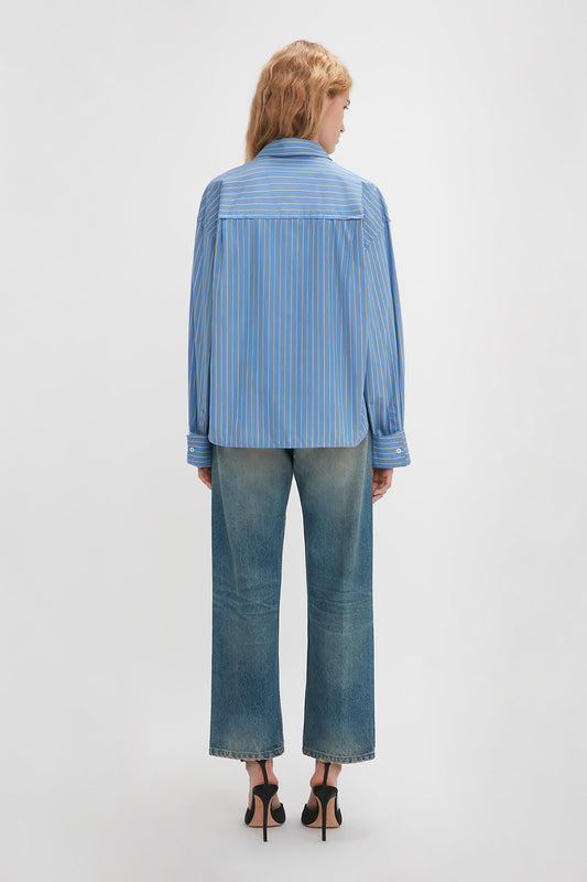 A person with blond hair is standing and facing away, wearing a blue pinstripe Cropped Seam Detail Shirt In Steel Blue by Victoria Beckham, high-rise wide-leg jeans, and black heels against a plain white background.