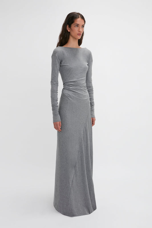 A woman with long dark hair is wearing a Victoria Beckham Long Sleeve Circle Neck Dress In Grey Marl, floor-length and gray, tailored from a super-soft cotton-jersey blend. She stands gracefully in front of a plain white background.