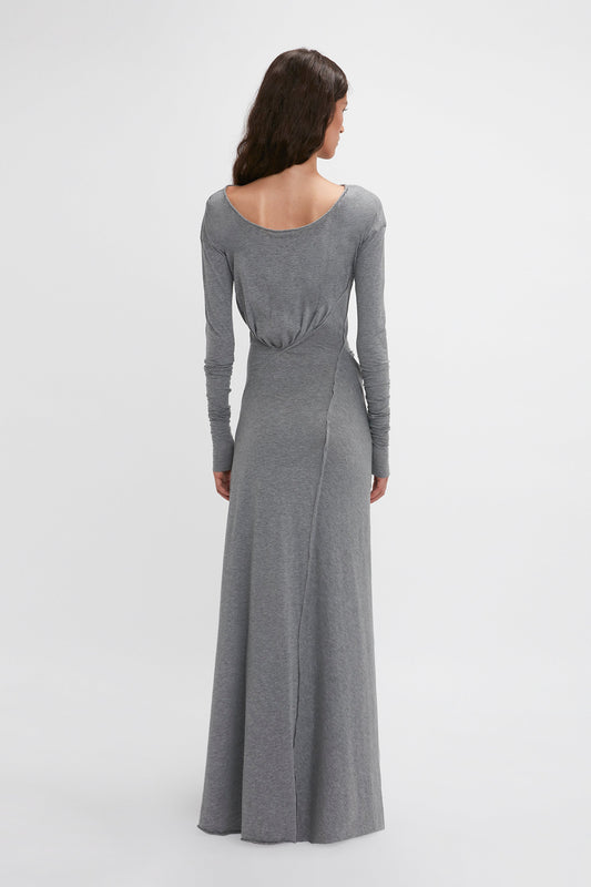 A person with long hair stands facing away, wearing an elegant Long Sleeve Circle Neck Dress In Grey Marl by Victoria Beckham in a super-soft cotton-jersey blend, featuring a scoop back and floor-length hem, epitomizing contemporary eveningwear.
