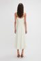 A woman in an Exclusive Panelled Midi Cami Dress In Vanilla by Victoria Beckham stands facing away from the camera. She has long, wavy brown hair and is wearing high-heeled shoes.