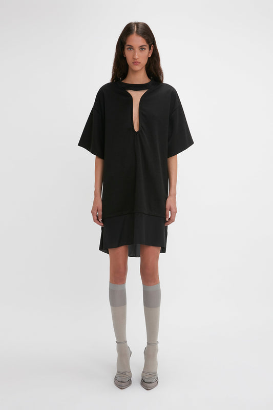 A woman stands against a plain background wearing the Victoria Beckham Frame Cut-Out T-Shirt Dress In Black, grey knee-high socks, and pointed-toe shoes, resembling an ensemble straight off the SS24 runway.