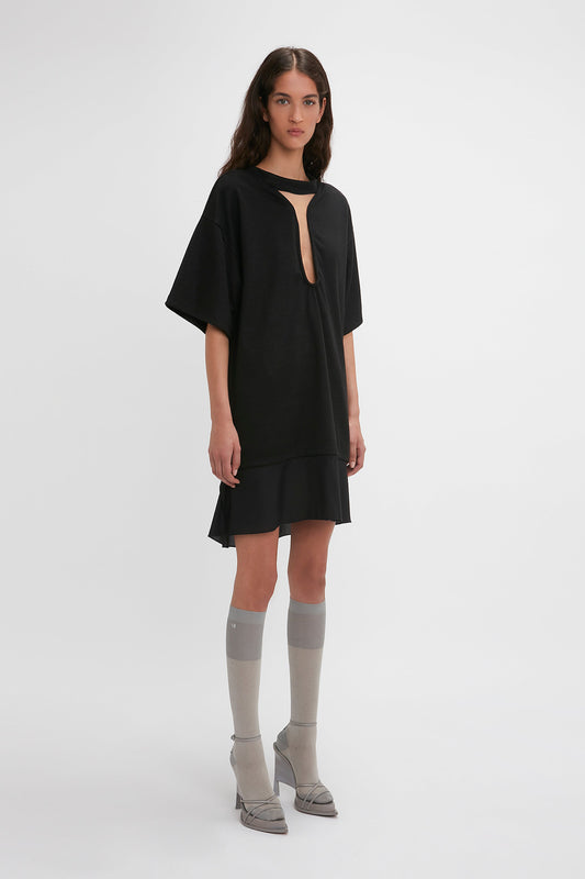 A woman with long hair wearing a loose-fitting black Frame Cut-Out T-Shirt Dress In Black by Victoria Beckham, paired with grey knee-high socks and grey shoes. She stands against a plain white background, embodying the effortless style of the SS24 runway.