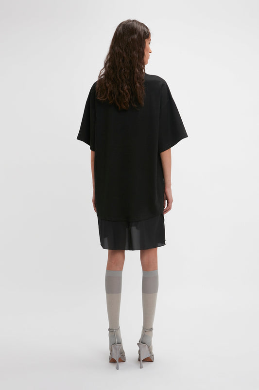 A person with long, wavy hair is standing, facing away, wearing a black oversized short-sleeve top that resembles a deconstructed Frame Cut-Out T-Shirt Dress In Black from Victoria Beckham, a black skirt, tall gray socks, and gray high-heeled shoes. The background is plain white.