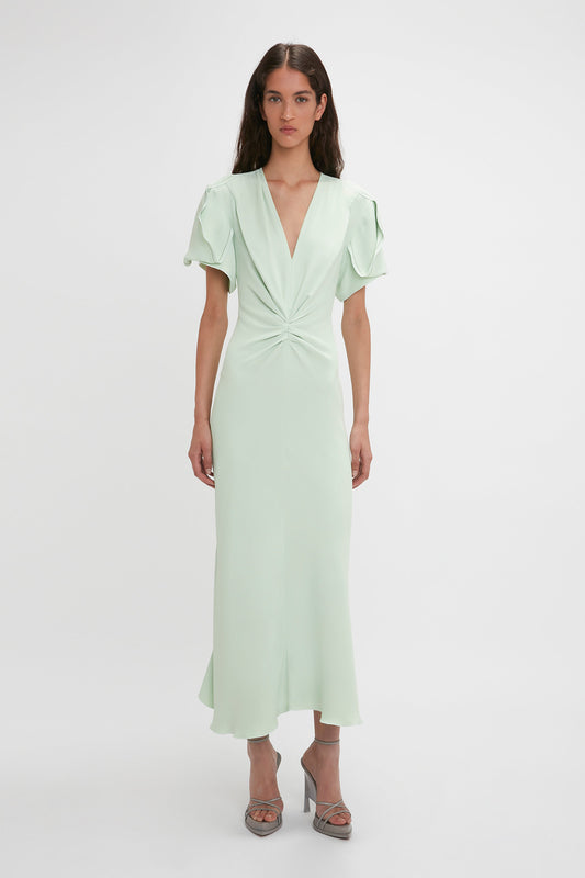 A woman stands posing in a light green, Victoria Beckham Gathered V-Neck Midi Dress In Jade with short sleeves and waist-defining pleat detail, paired with silver heels, on a white background.