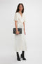 A woman in a Gathered V-Neck Midi Dress In Ivory by Victoria Beckham with waist-defining pleat detail and draped sleeves, paired with black heels, holds a black clutch with gold lettering, standing against a plain white background.