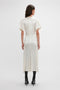 A person with long hair stands facing away, wearing a Victoria Beckham Gathered V-Neck Midi Dress In Ivory featuring elegant waist-defining pleat detail and draped sleeves, paired with black heeled boots, against a plain white background.