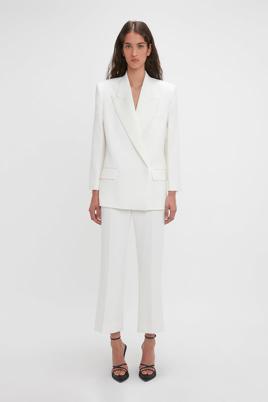A woman stands wearing a white double-breasted blazer and matching Victoria Beckham Exclusive Cropped Tuxedo Trouser In Ivory, paired with black high-heeled shoes, against a plain white background.