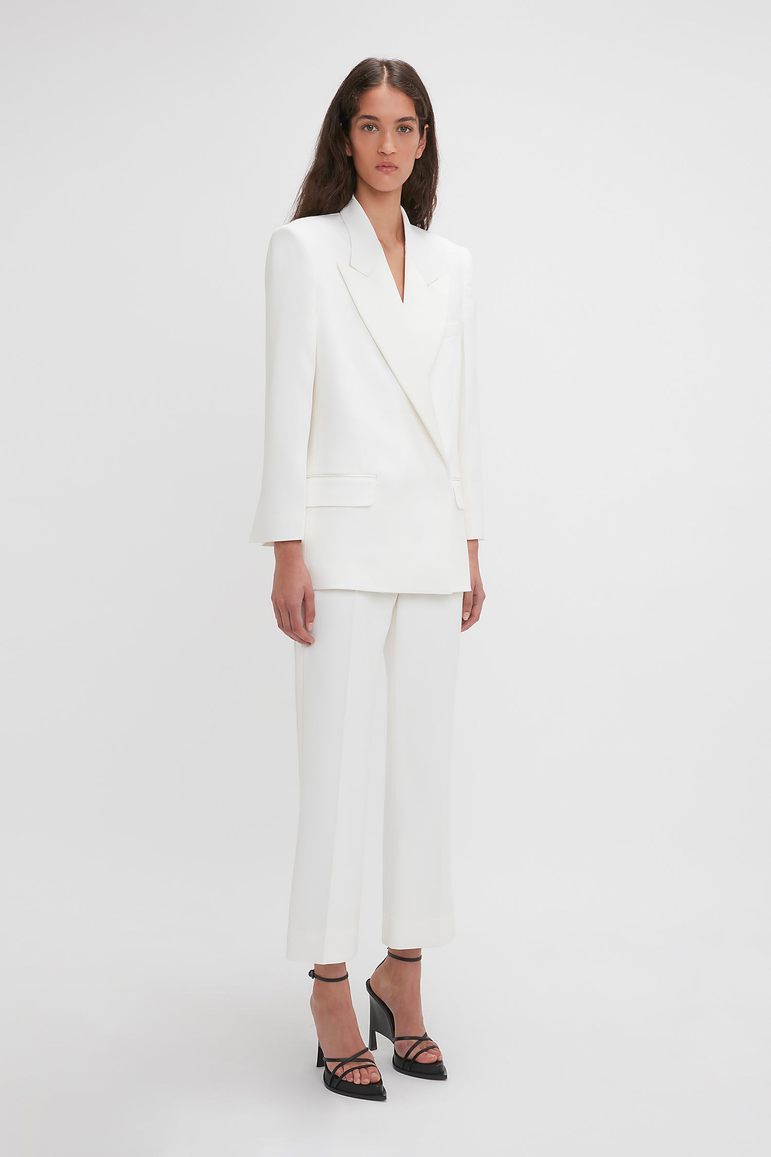 A woman is standing against a white background, wearing an Exclusive Double Breasted Tuxedo Jacket In Ivory by Victoria Beckham with matching trousers, complemented by black strappy high-heel shoes—perfect for black-tie occasions.