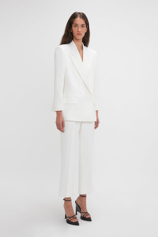 A woman is standing against a white background, wearing an Exclusive Double Breasted Tuxedo Jacket In Ivory by Victoria Beckham with matching trousers, complemented by black strappy high-heel shoes—perfect for black-tie occasions.
