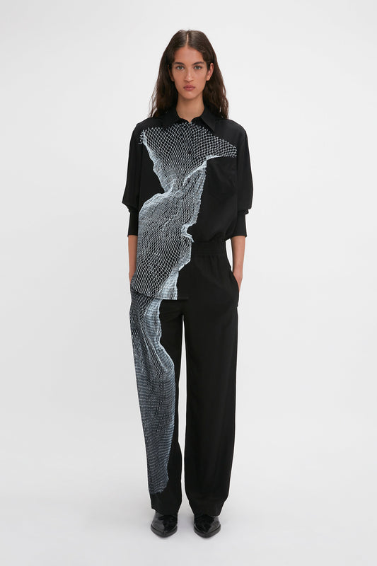 A person stands against a white background, wearing wide leg Pyjama Trousers In Black-White Contorted Net from Victoria Beckham and a black shirt with a distinctive twisted net print on the outfit’s left side. They have long dark hair and are looking straight ahead.