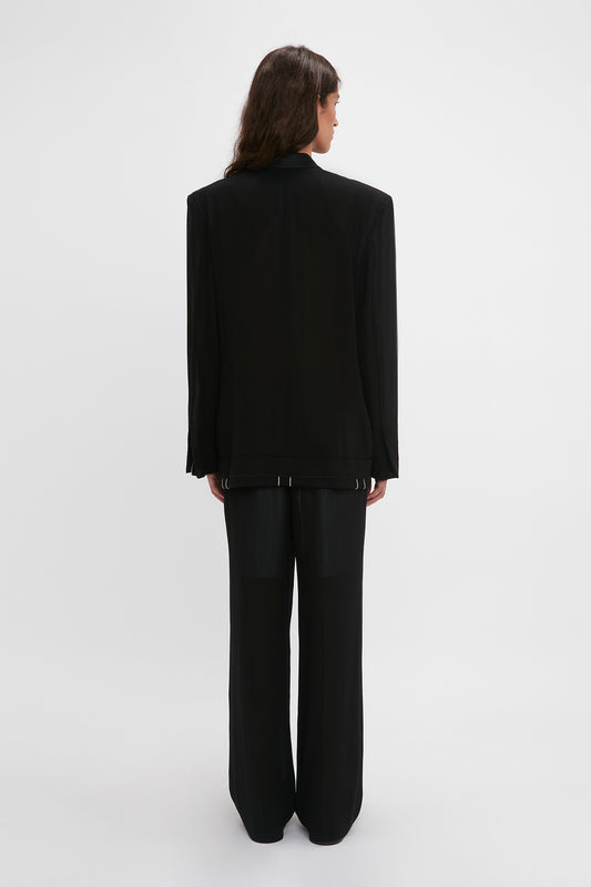 Woman standing, viewed from behind, wearing a Victoria Beckham black wool tailored jacket with folded detail and straight leg trousers on a white background.