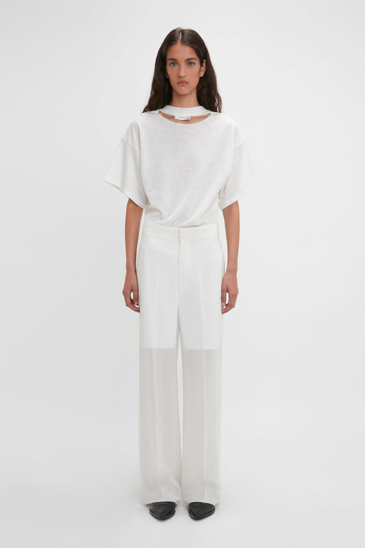 Woman wearing a white short-sleeve top with cut-out detail at the neckline and Waistband Detail Straight Leg Trouser In White by Victoria Beckham, standing against a plain white background. She is also wearing black flat shoes.