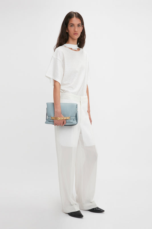 A woman in a white t-shirt and sheer white pants holds a Victoria Beckham Puffy Chain Pouch With Strap In Ice Leather, standing against a plain background.