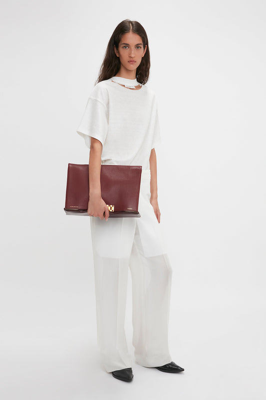 A woman stands holding a large burgundy clutch. She is wearing a white short-sleeve top, Waistband Detail Straight Leg Trouser In White by Victoria Beckham, and black shoes. The background is plain white, highlighting her contemporary fashion sense.