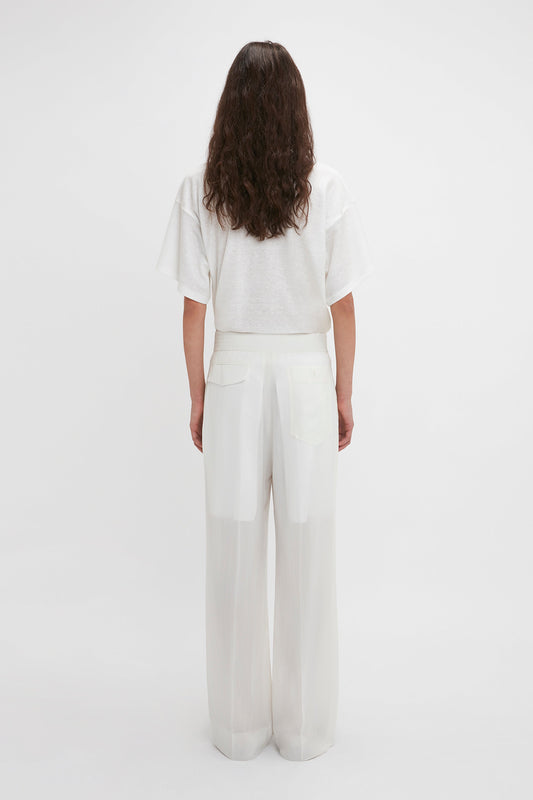 A person with long hair is standing with their back to the camera, wearing a white T-shirt and contemporary fashion Waistband Detail Straight Leg Trouser In White by Victoria Beckham.