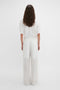 A person with long hair is standing with their back to the camera, wearing a white T-shirt and contemporary fashion Waistband Detail Straight Leg Trouser In White by Victoria Beckham.