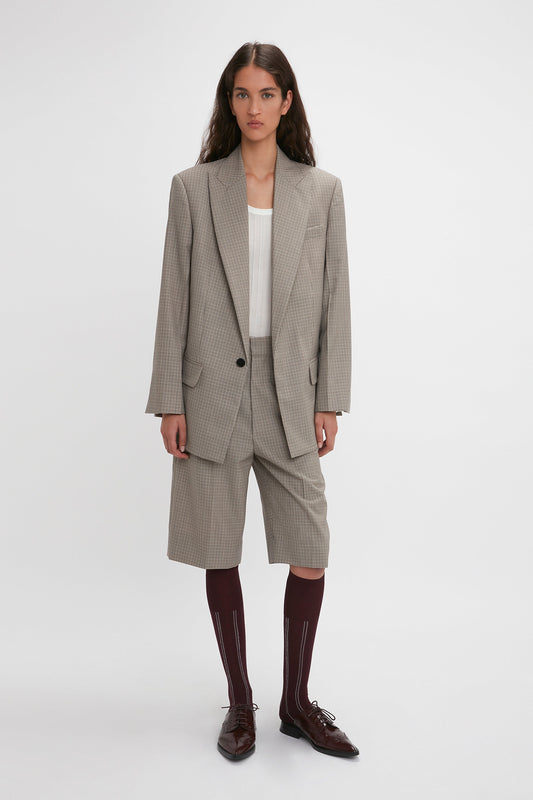 A person with long hair stands against a plain background wearing the Victoria Beckham Peak Lapel Jacket In Multi with a contemporary aesthetic, matching shorts, a white top, maroon knee-high socks, and brown dress shoes.