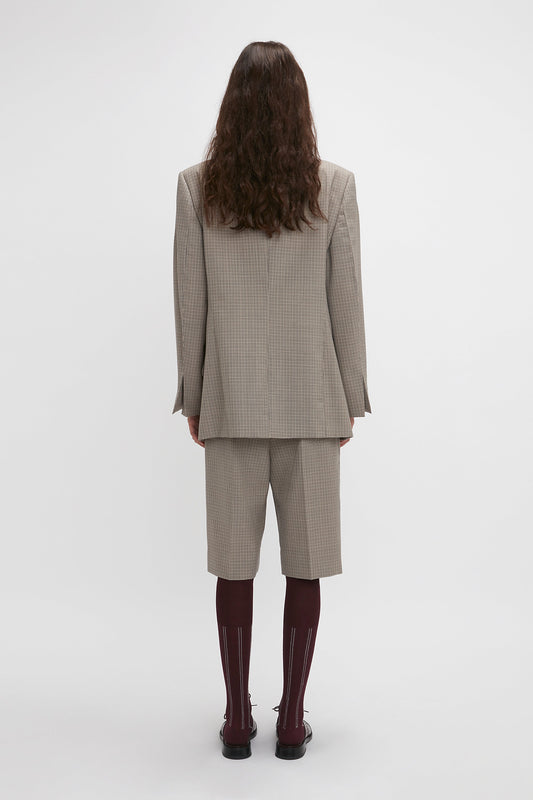 A person with long hair is standing facing away, wearing a beige plaid Peak Lapel Jacket In Multi by Victoria Beckham, matching shorts, burgundy knee-high socks, and black shoes, creating a contemporary aesthetic.