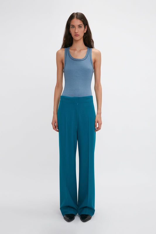 A woman with long hair is standing against a plain background, wearing a blue sleeveless top and Waistband Detail Straight Leg Trouser In Petroleum by Victoria Beckham featuring precision tailoring.