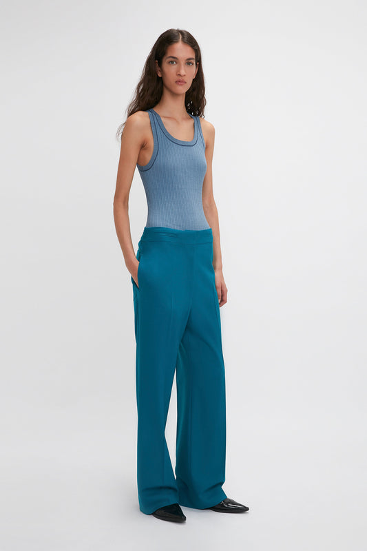 A woman stands against a plain background, clad in a blue sleeveless top and Victoria Beckham Waistband Detail Straight Leg Trouser In Petroleum. Her hands rest in her pockets as she faces the camera, showcasing the precision tailoring of her outfit.