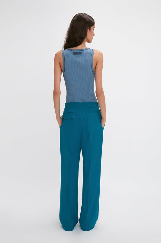 A person with long hair wearing a blue sleeveless top and Waistband Detail Straight Leg Trouser In Petroleum by Victoria Beckham, known for their precision tailoring, is standing and facing away from the camera.