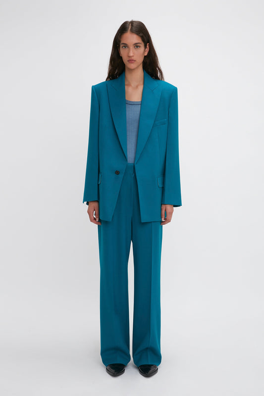A woman stands against a plain background wearing an oversized Peak Lapel Jacket In Petroleum and matching wide-leg trousers over a light blue top, showcasing precision tailoring reminiscent of Victoria Beckham's style.
