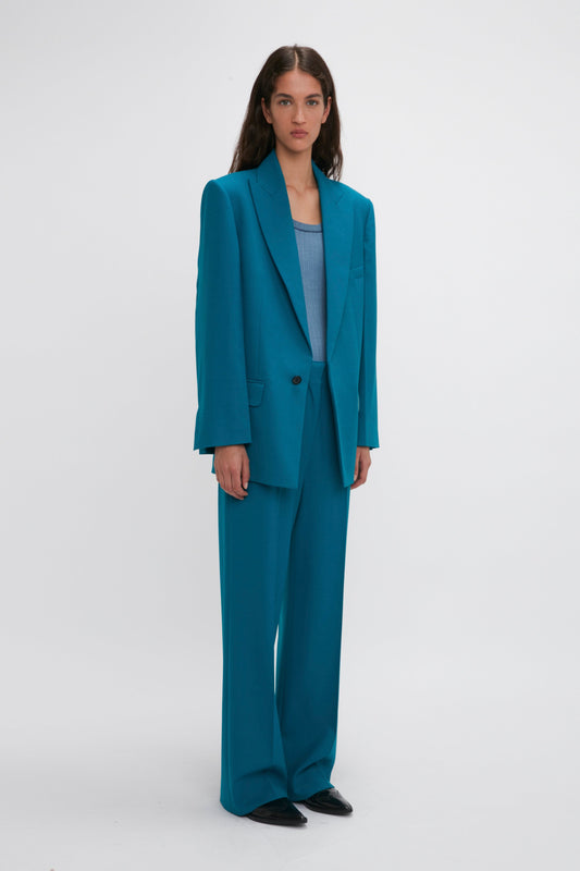 A woman stands against a plain white background, wearing an oversized teal suit with precision tailoring inspired by Victoria Beckham. The ensemble includes wide-leg trousers, a lighter blue top, and sharp black shoes, all accentuated by the Peak Lapel Jacket In Petroleum from Victoria Beckham.