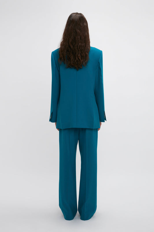 Person with long, curly hair wearing a teal pantsuit featuring precision tailoring and a Peak Lapel Jacket In Petroleum by Victoria Beckham, viewed from the back against a plain white background.