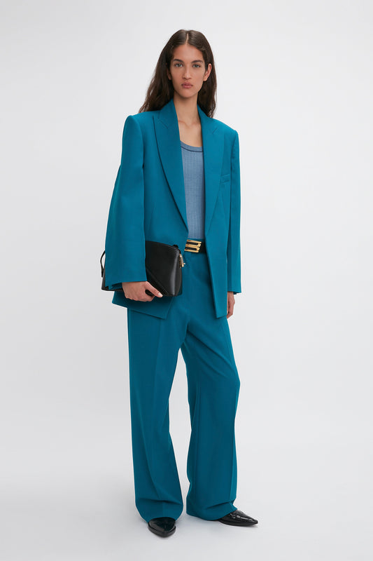 A person stands against a plain background, wearing a teal Victoria Beckham suit with an oversized Peak Lapel Jacket In Petroleum featuring a peak lapel jacket and wide-leg trousers, holding a black clutch and wearing a blue top.