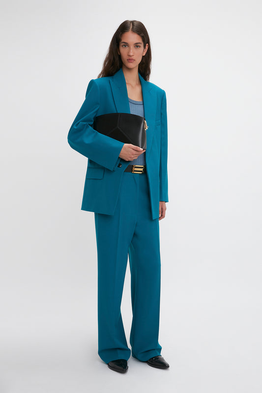A woman stands against a plain background wearing a teal Waistband Detail Straight Leg Trouser In Petroleum by Victoria Beckham, holding a black clutch bag. She has long dark hair and is looking directly at the camera.