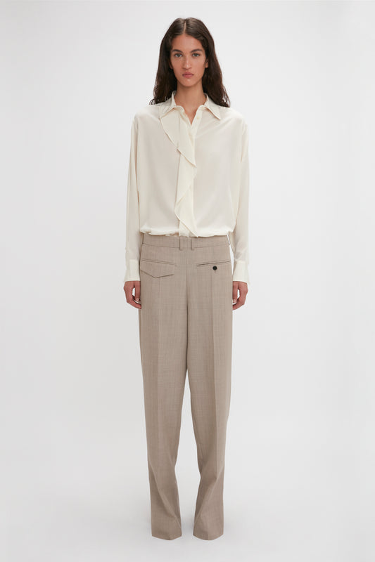 A person stands against a plain background, wearing the Victoria Beckham Asymmetric Ruffle Blouse In Ivory with voluminous sleeves and a ruffled front, paired with beige trousers featuring a button-front closure.